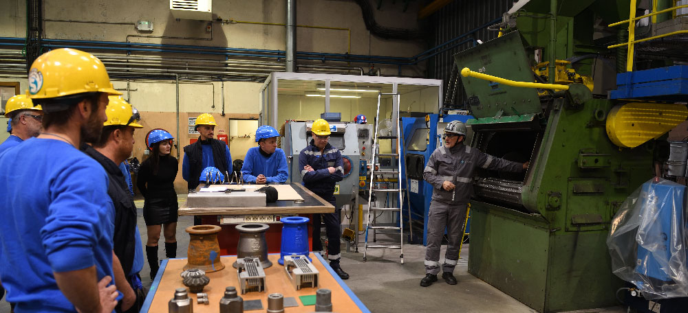 This image shows a group of professionals participating in a training session for shot blasting (grenaillage). They are gathered around an industrial tumblast machine, part of Winoa's training program. The group, wearing safety gear including hard hats and work uniforms, is being guided by an expert who appears to be explaining how the shot blasting machine works. Various shot blasting components are displayed on a table in front of the group, showcasing tools and equipment used in the process. The environment is a workshop setting, within Winoa France technical center .